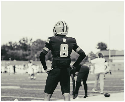 A person wearing a football uniform with the number eight on the back stands on a field with hands on hips while other people and players are visible in the background. Description generated by AI