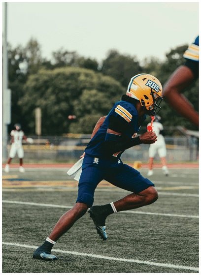 An adult wearing a yellow helmet and blue football uniform is running on a football field while holding a football. Description generated by AI