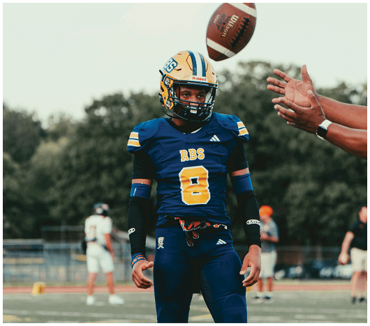 A person in a blue and yellow football uniform with the number 8 stands on a field while a football is being thrown toward them. Description generated by AI