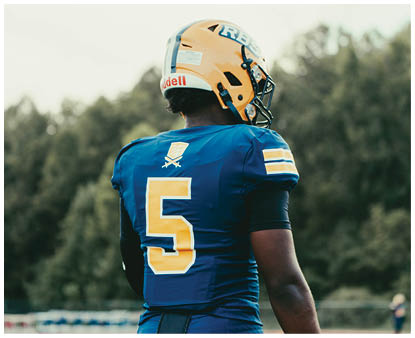 A person wearing a blue football jersey with the number five and a yellow helmet is standing outdoors with trees in the background. Description generated by AI