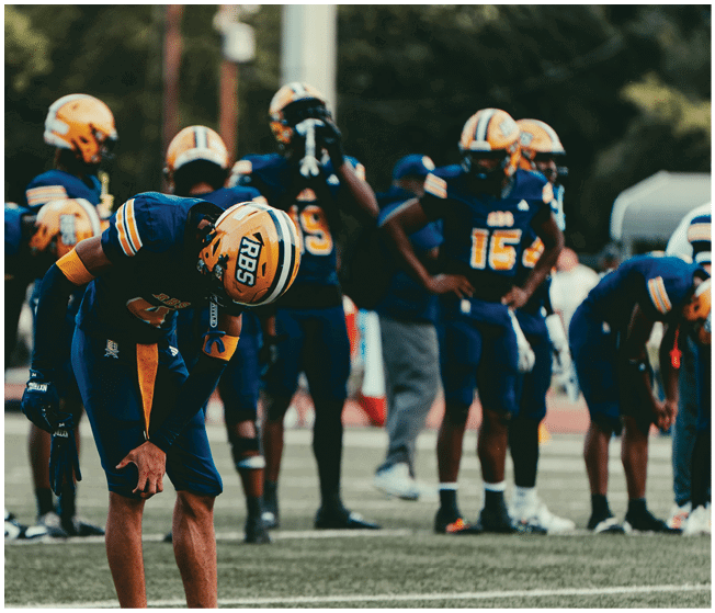 People wearing blue and orange football uniforms are gathered on a field, with one person bending forward in the foreground and others standing or drinking water in the background. Description generated by AI
