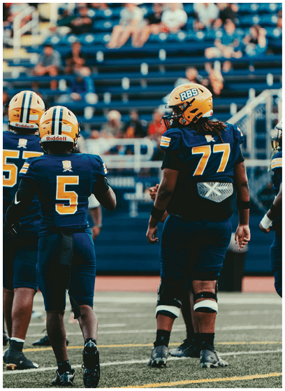 Several people wearing dark blue football uniforms and yellow helmets stand on a field with bleachers and spectators in the background. Description generated by AI