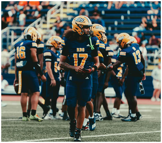 A group of people wearing blue and yellow football uniforms with helmets are gathered on a field with blurred spectators in the background. Description generated by AI