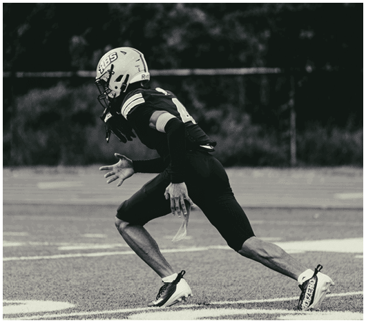 A person wearing a football helmet and uniform is running on a field with a blurred background of trees. Description generated by AI
