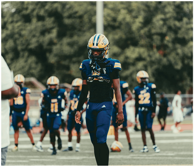 A person in a blue and yellow football uniform is walking on a field with several other similarly dressed people in the background. Description generated by AI