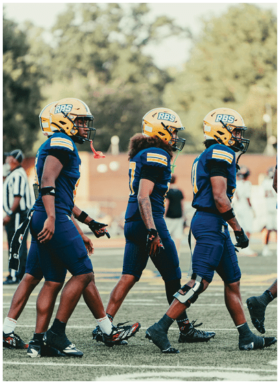 Three adults in blue and yellow football uniforms and helmets walk side by side on a field with blurred trees and people in the background. Description generated by AI