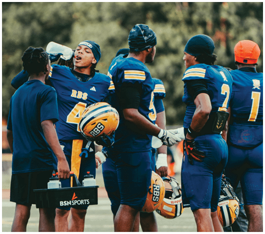 People in blue football uniforms are gathered on a field with one person drinking water and another holding a sports equipment case. Description generated by AI