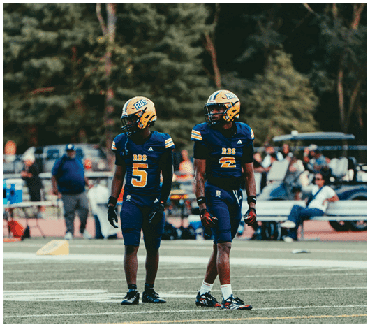 Two people wearing blue and yellow football uniforms and helmets stand on a football field with trees and spectators in the background. Description generated by AI