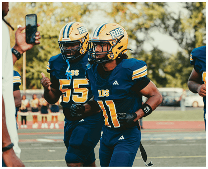 Two people in blue and yellow football uniforms are running on a field during a game. Description generated by AI