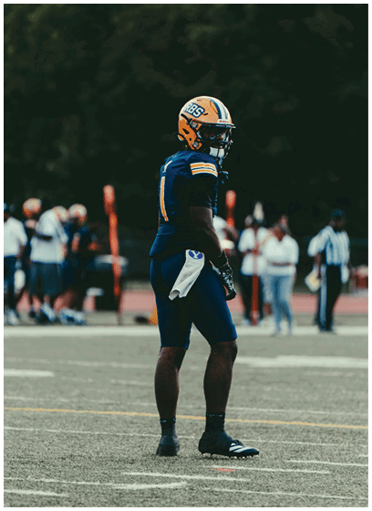 A person wearing a blue and yellow football uniform and helmet stands on a football field with blurred people and a referee in the background. Description generated by AI