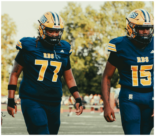 Two people wearing blue and yellow football uniforms with helmets are walking on a field. Description generated by AI