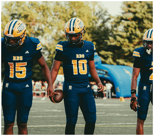Three people in football uniforms stand on a field holding hands, with one person holding a football. Description generated by AI