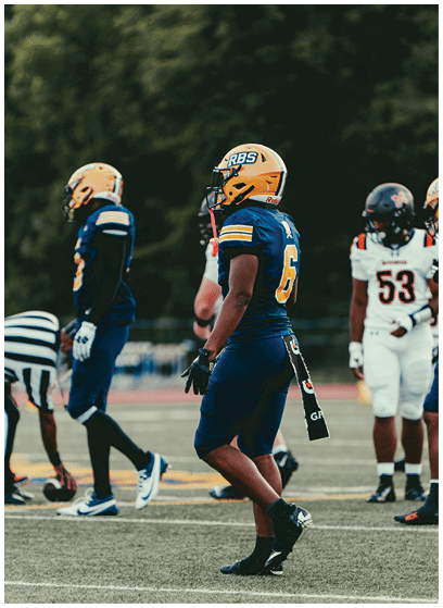 Several people wearing football uniforms and helmets stand on a field, with some preparing for a play and others observing. Description generated by AI