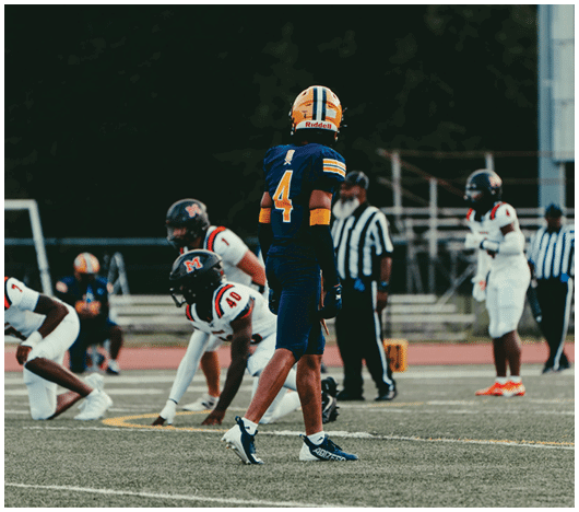 A group of people in football uniforms are on a field with one person in a blue and yellow uniform standing in the center while others in white uniforms prepare for a play. Description generated by AI