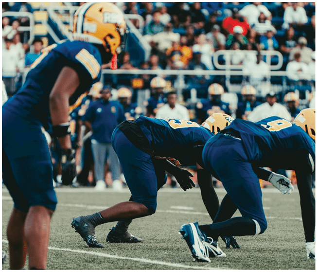 Three people in blue football uniforms and yellow helmets are crouched on a football field preparing for a play with a crowd visible in the background. Description generated by AI