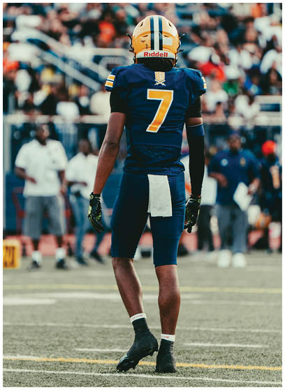 A person wearing a blue and yellow football uniform with the number seven on the back stands on a field facing away from the camera with a blurred crowd in the background. Description generated by AI
