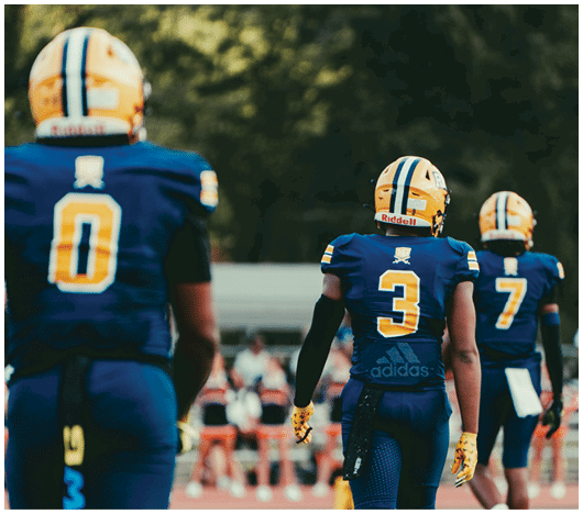 Three people wearing blue and yellow football uniforms with helmets are standing on a field with a blurred background of spectators. Description generated by AI