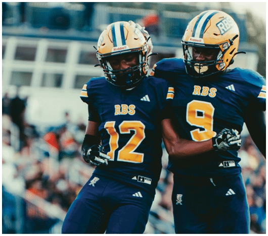 Two people in blue football uniforms with yellow numbers are on a field, with one person mid-jump and a blurred crowd in the background. Description generated by AI