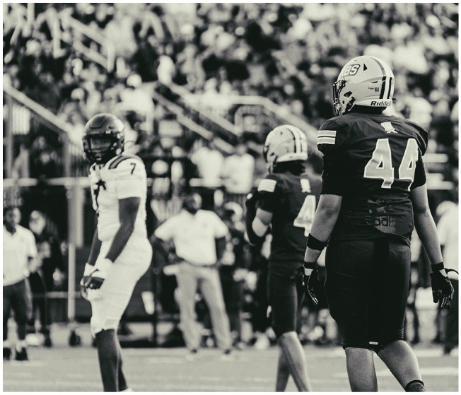 Three people in football uniforms stand on a field with a blurred crowd in the background, with one person in a dark uniform facing away and two others in lighter uniforms facing different directions. Description generated by AI