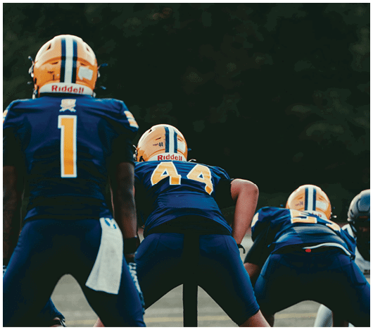 Two people wearing blue football uniforms and yellow helmets are positioned on a field, preparing for a play. Description generated by AI