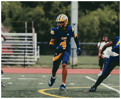 An adult wearing a blue and yellow football uniform with the number four is running on a football field with other players and a blurred background of bleachers and trees. Description generated by AI