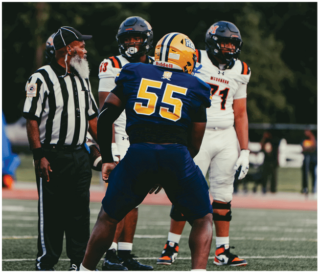 A referee and three football players in uniforms are standing on a field during a game, with two players in white and one in blue facing the referee. Description generated by AI