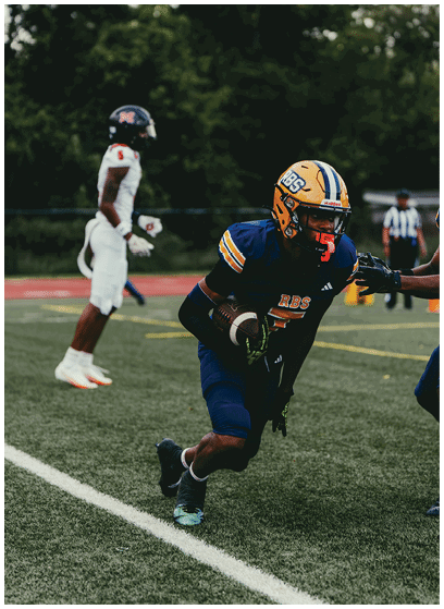A person in a blue and yellow football uniform is running on a field while holding a football, with another person in a white uniform visible in the background. Description generated by AI