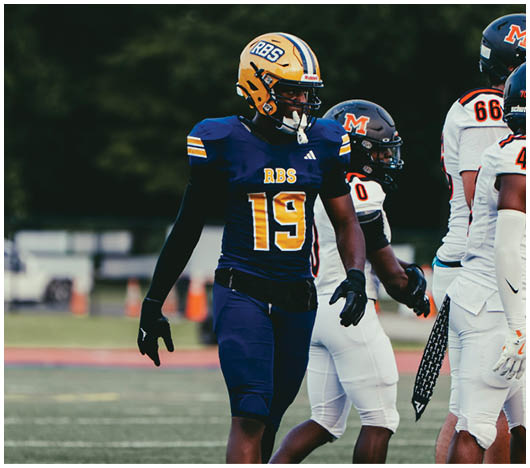 A person wearing a blue and yellow football uniform with the number 19 and helmet labeled RBS is standing on a field near other players in white and black uniforms. Description generated by AI