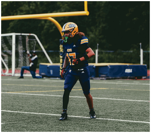 A person wearing a blue and yellow football uniform and helmet is walking on a field near a goalpost and soccer net. Description generated by AI