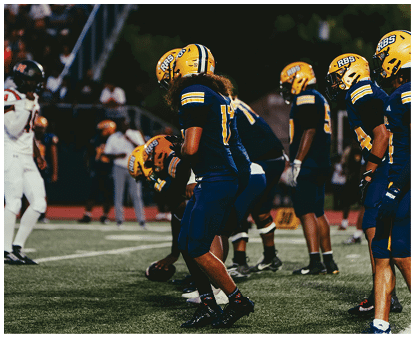 A group of people wearing yellow helmets and dark blue sports uniforms are lined up on a football field preparing for a play against opponents in white uniforms. Description generated by AI