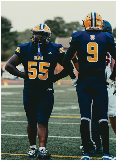 Two people in football uniforms with helmets stand on a field, one facing forward with number 55 and the other facing away with number 9. Description generated by AI