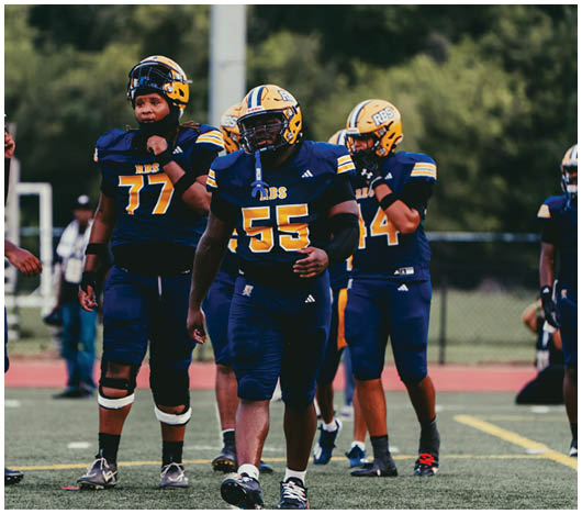 Four people in blue and yellow football uniforms stand on a field with one person walking toward the camera and others standing behind. Description generated by AI