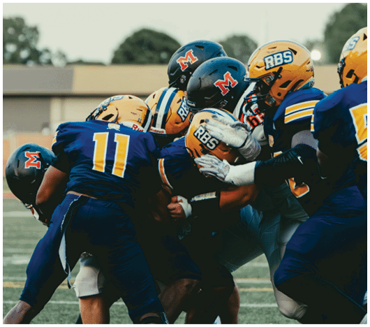 Several people in football uniforms are engaged in a close tackle on a grassy field during a game. Description generated by AI