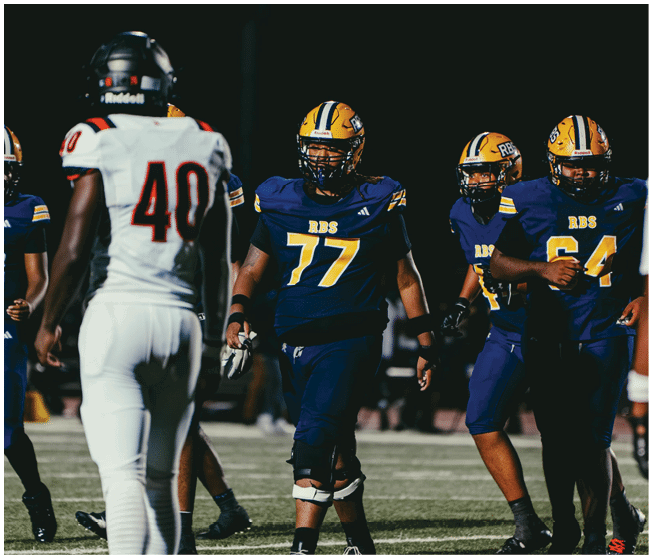 Several people wearing football uniforms are on a field, with some in dark blue and yellow jerseys and one in a white jersey. Description generated by AI