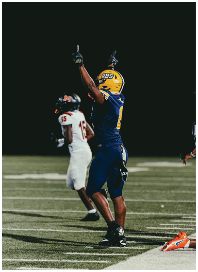 A person in a blue and yellow football uniform is raising their arms on a football field while another person in a white uniform with the number 15 is in the background. Description generated by AI