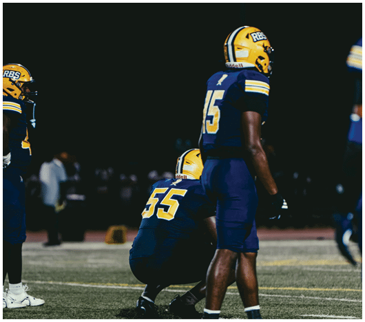 Two people wearing football uniforms and helmets are on a field, with one person squatting and the other standing. Description generated by AI