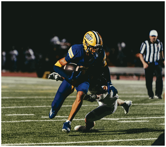 A person in a blue and yellow football uniform is running with the ball while another person in a white uniform attempts to tackle them on a football field at night. Description generated by AI