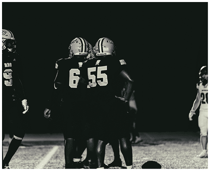 Four people in football uniforms stand on a field at night, with three grouped closely together and one standing apart on the right. Description generated by AI