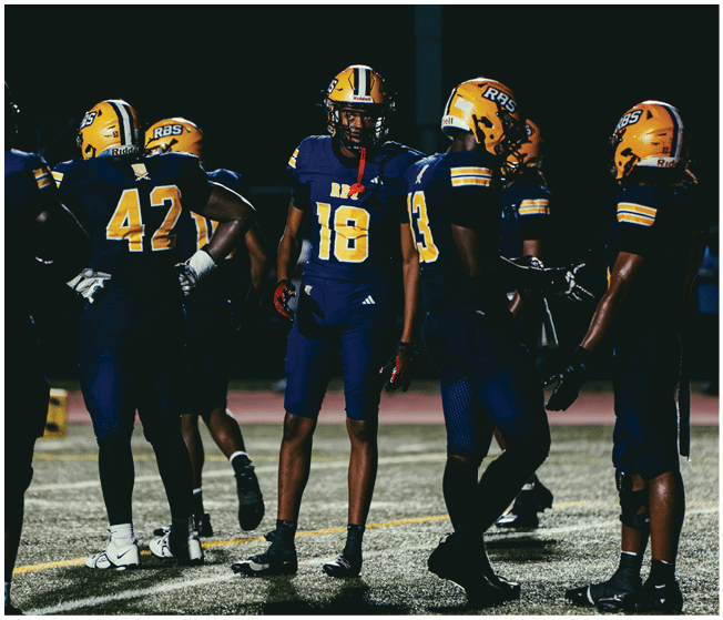 A group of people wearing yellow helmets and dark blue football uniforms are standing on a field at night. Description generated by AI