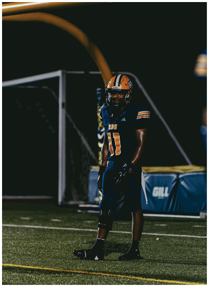A person wearing a dark blue and orange football uniform with the number 11 stands on a football field at night. Description generated by AI