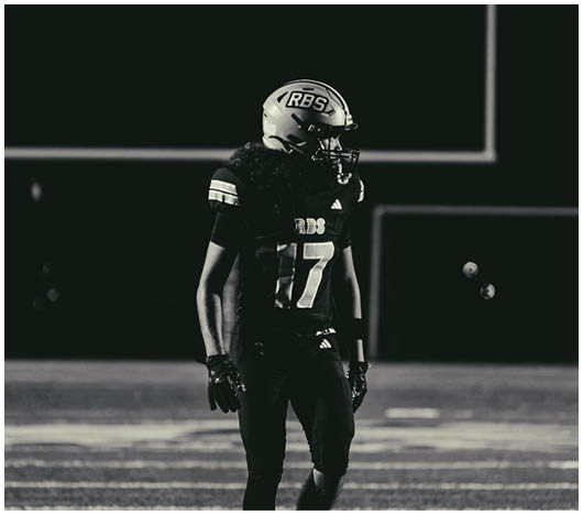 A person wearing a football uniform and helmet stands on a field at night with a goalpost visible in the background. Description generated by AI