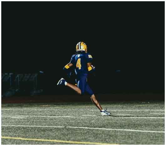 A person wearing a blue and yellow football uniform with the number four is running on a field at night. Description generated by AI