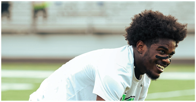 A person with curly hair and black face paint under their eyes is smiling and leaning forward on a sports field. Description generated by AI
