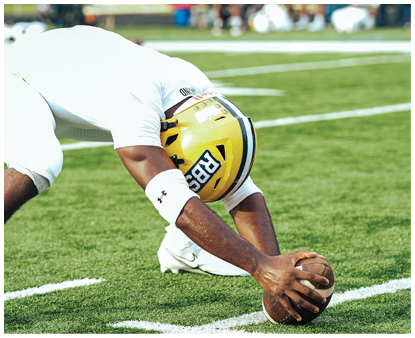 A person in white athletic gear and a yellow helmet is bent over on a grassy sports field with blurred people and benches in the background. Description generated by AI