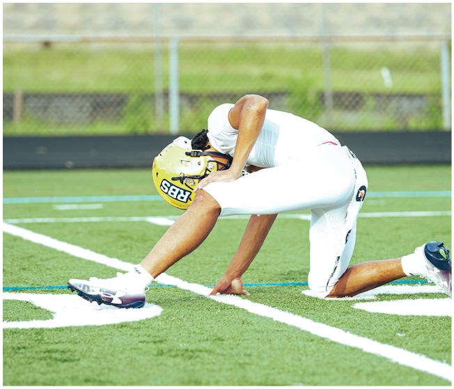 A person wearing a yellow helmet and white football uniform is stretching on a green football field near white yard lines. Description generated by AI