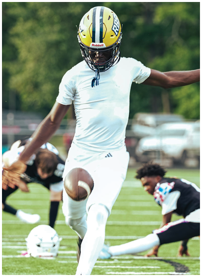 A person wearing a yellow helmet and white athletic clothing is kicking a football on a grassy field while another person in black and white sportswear is kneeling in the background. Description generated by AI