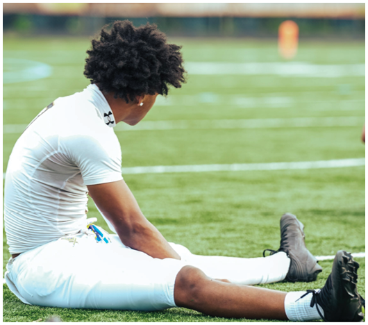 A person with curly hair wearing a white athletic outfit is sitting on a grassy sports field stretching their legs. Description generated by AI