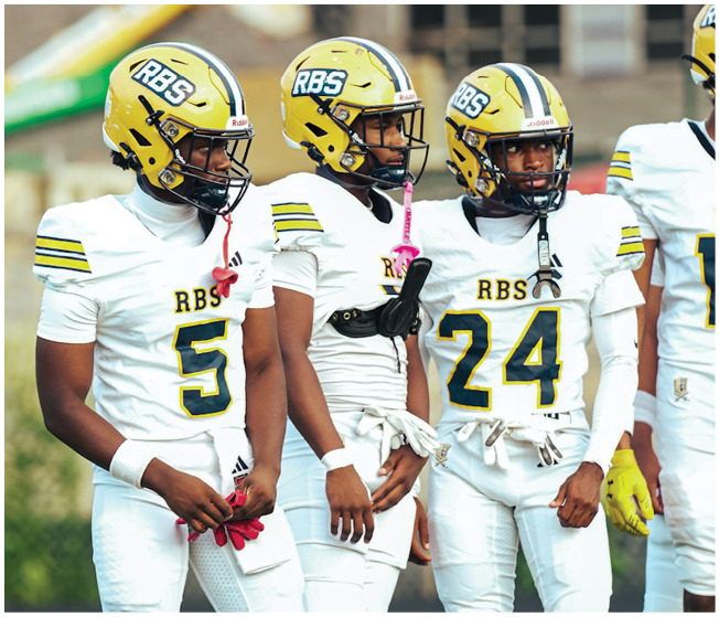 Three adults wearing white football uniforms and yellow helmets stand on a field near a lacrosse goal. Description generated by AI