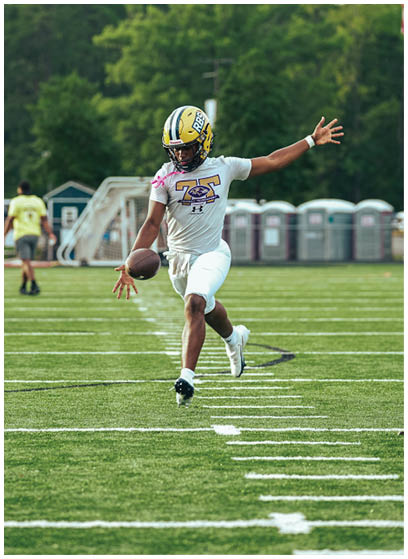 A person wearing a football helmet and uniform is running on a football field while holding a football, with portable toilets and trees visible in the background. Description generated by AI