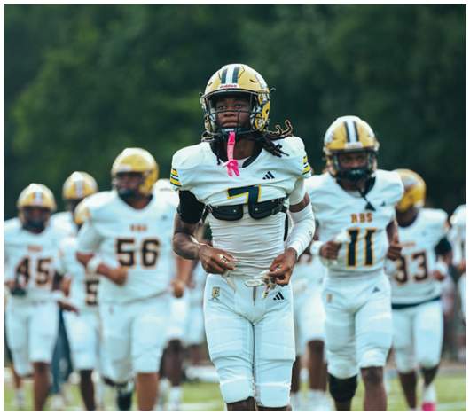 A group of people in white and gold football uniforms are running on a field with one person wearing number 7 leading the group. Description generated by AI